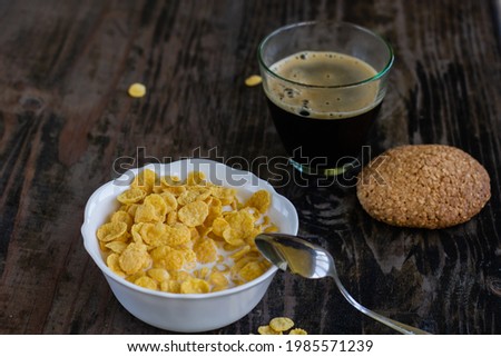 A white plate with yellow corn cereal healthy sugar-free flakes with milk a glass of coffee or tea stands on a brown table next to oatmeal cookies