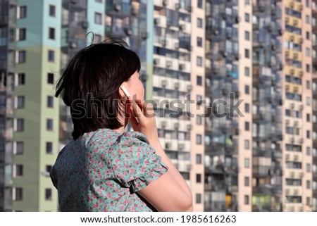 Woman touches wireless earphone in the ear standing on residential buildings background. Headset, listening to music and voice call concept