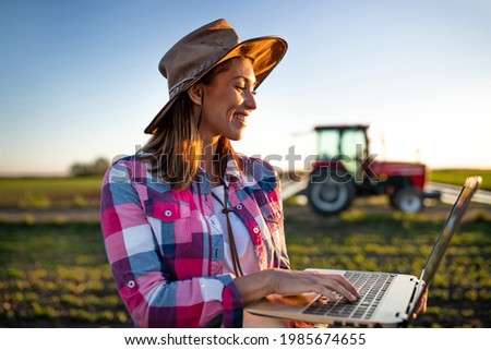 Female agronomist using laptop modern technology in agriculture. Young farmer standing in field in front of tractor machinery smiling. 