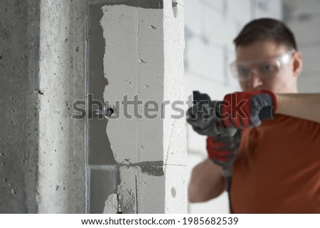 A builder with glasses drills a hole in the wall to see through. The master man makes a hole in the wall with a percussion drill. Construction drill drill for concrete creating a hole. Royalty-Free Stock Photo #1985682539