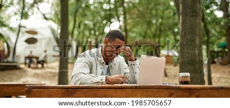 Happy young african american man freelancer working on laptop while sitting at wooden table outdoors, panorama. Work in nature concept