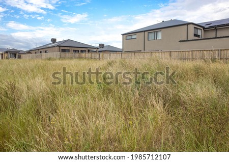 Vacant land block covered with long grass, some modern residential houses in the background. Concept of real estate development, and land for sale. Tarneit, Melbourne, VIC Australia. Royalty-Free Stock Photo #1985712107
