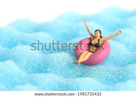 Miniature people toy figure photography. Girl wearing black sunglass swimming with rubber tube ring on wavy ocean. Image photo Royalty-Free Stock Photo #1985735432