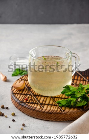 Chicken broth in a glass cup with parsley, garlic and other spices on a wooden board on a gray concrete background. Copy space