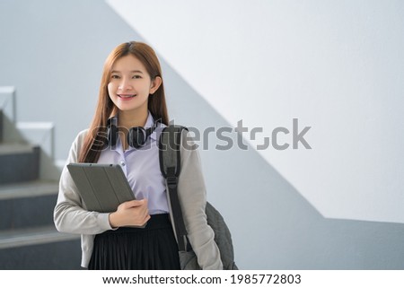 ํYoung Asian woman student in uniform holding a tablet in smart and happy pose in university or college classroom. Youth girl student and tutoring education with technological learning concept