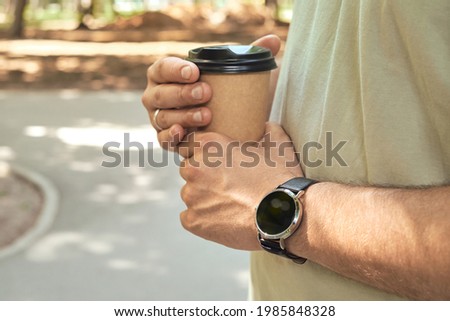 Craft paper cup with coffee in the hand of a man with a clock. Disposable paper cup close up. A man checks the time on his stylish watch. Time management, mobility, social and technological concept