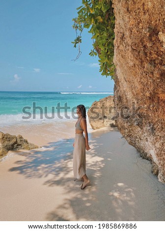 European girl wearing swimsuit and beige culottes standing on a golden tropical sandy and rocky Bali beach with turquoise ocean water background