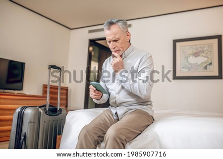 Mid-aged businessman sitting on a bed in a hotel with a smartphone in hands