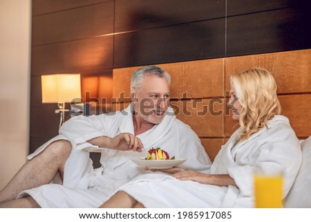 A wife and a husband eating their breakfast in bed and enjoying it