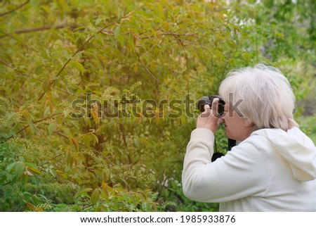 Senior woman shooting photo by digital camera. Shooting blooming bird cherry tree. Concept of aged people and photography. Cloudy weather