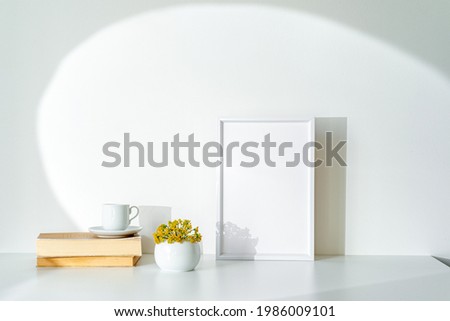 Empty white picture frame mockup in sunlight. Books, coffee cup and sphere-shaped vase with small yellow flowers. Concept of waking up and good morning with optimism. White table and wall 