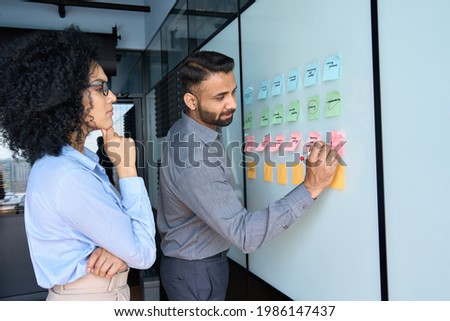 Young indian businessman manager writing strategy ideas on sticky notes on whiteboard and female African American colleague looking at his strategy scrum presentation. Business project planning. Royalty-Free Stock Photo #1986147437