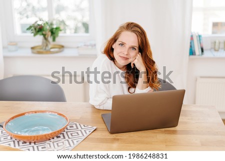 Smiling friendly young redhead woman leaning her chin on her hand looking attentively at the camera as she sits working at a laptop computer at home