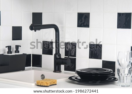 on the table, against the background of black and white tiles, there is a stack of dishes, cleaning agent, there is a sink with a tap