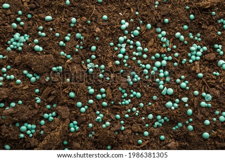 Green complex fertiliser granules on dark brown soil. Closeup. Product for root feeding of vegetables, flowers and plants. Royalty-Free Stock Photo #1986381305