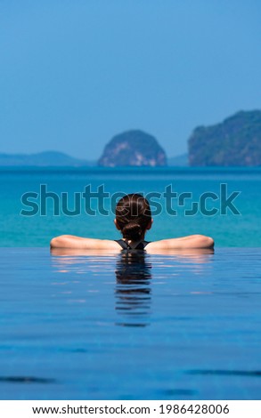 portrait of young woman wearing bikini standing in the blue infinity swimming pool looking at the view of ocian on summer vacation
