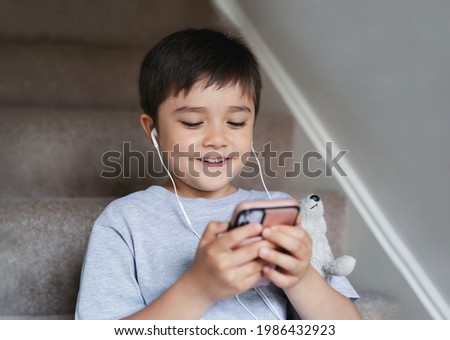 Candid shot child sitting on staircase wearing earphones listening relaxing music at home.Portrait happy boy playing games on mobile phone, Kid watching cartoon on cell phone.Children and Technology 