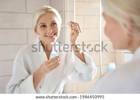 Senior mature older caucasian woman touching clean face eye contour with antiaging pipette serum essence oil looking at mirror wearing bathrobe. Anti wrinkle prevention skin care products concept. Royalty-Free Stock Photo #1986450995