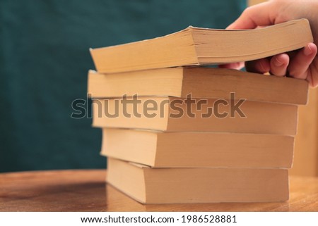 Hand putting book on books. Books on a wooden table. A stack of book on petrol green background with copy space for text. Educational or novel books. Back to school background. Royalty-Free Stock Photo #1986528881