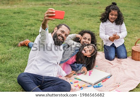 Happy indian family enjoy day outdoor at city park with pic nic and toys while taking a selfie with smartphone - Family, parents and children love