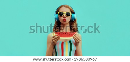 Summer portrait of stylish woman in headphones listening to music blowing her lips sends kiss with fresh juicy slice of watermelon on blue background, blank copy space for advertising text