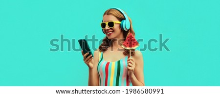 Summer portrait of happy smiling young woman model in headphones listening to music on smartphone with juicy lollipop or ice cream shaped slice of watermelon on blue background