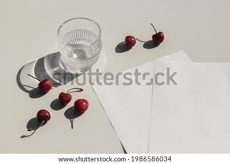 Summer food still life composition. Fresh red cherries fruit on beige table background. Stationery mock up scene. Blank paper pages. Glass of water, cocktail glittering in sunlight. Top view. Royalty-Free Stock Photo #1986586034