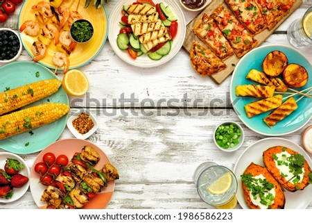 Summer BBQ grill frame over a white wood background. Chicken and shrimp skewers, flatbread, stuffed sweet potato, grilled fruit, corn and salad. Top down view with copy space.