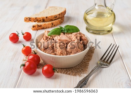Canned tuna meat in a bowl, fork, bread and fresh red cherry tomatoes on a white wooden table. Low calories healthy eating snack of preserved tuna fish and vegetables. Tasty seafood. Front view. Royalty-Free Stock Photo #1986596063
