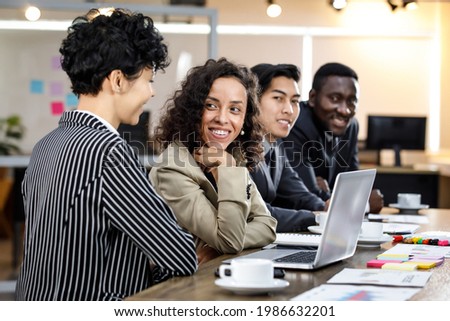 Close up to two mixed race caucasian african beautiful smart professional businesswomen talking, discussing job strategy, using laptop for working in indoor office with background of male colleagues