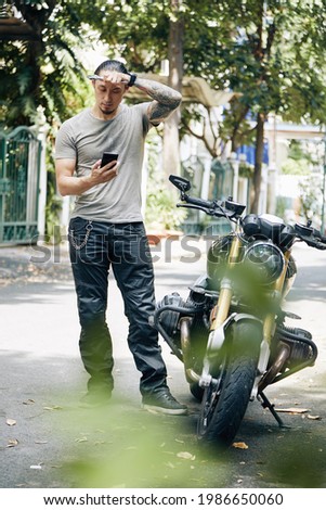Confused man with wrench in hands wiping off sweat from her forehead and reading article on motorcycle repairing on his smartphone