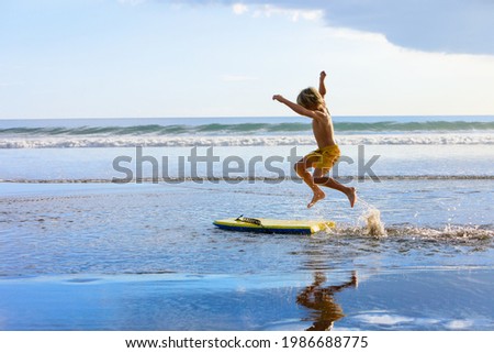 Happy boy - young surfer with bodyboard have fun on beach, run by sea water pool. Active family lifestyle, kids outdoor water sports, swimming activity in surf camp. Summer vacation with child.