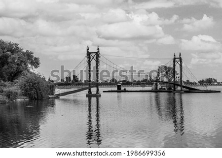 Spring scenery of a reflective surface of a lake, cloudy sky and distant bridge with line of trees. Monochrome picture