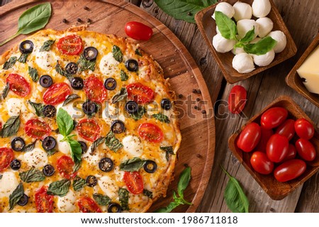 On a wooden stand, we see a beautifully decorated pizza. To the right of the pizza, in wooden bowls, are tomatoes, mushrooms and fresh green mint leaves. View from above.