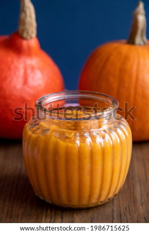 Side view of open glass jar with pumpkin puree standing by pumpkin vegetable on wooden table. Close-up view. Selective focus. Organic food theme.