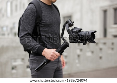 videography, filmmaking and creativity concept - close up of modern dslr camera on 3-axis gimbal stabilizer in male hands over concrete building background Royalty-Free Stock Photo #1986724094