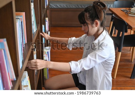 Female college student sitting near bookshelves and looking for a book in the library.