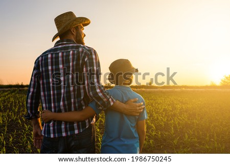 Father and son are standing in their growing wheat field. They are happy because of successful sowing and enjoying sunset. Royalty-Free Stock Photo #1986750245
