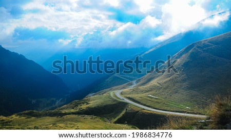 Sun shines through stormy clouds above Transfagarasan mountainous road. The lane winds under the sun rays through alpine pastures, above Capra Valley. Unique moments in Fagaras Massif, Carpathia. Royalty-Free Stock Photo #1986834776