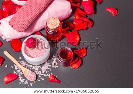 Natural spa concept with fresh roses petals. Rose sea salt, essential oil, soft towels. Modern hard light, dark shadow. Black stone concrete background, top view