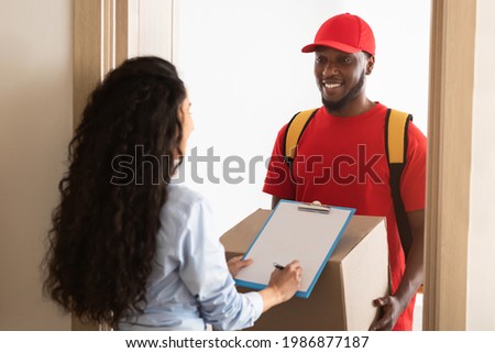 Fast Shipment Concept. Rear View Of Young Woman Receiving Order Signing Paper Form On Clipboard, Confirming Delivery. Cheerful African Postman In Red Uniform Standing At Door Holding Carton Box