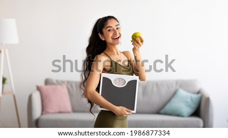 Portrait of millennial Indian lady holding scales and eating apple, choosing healthy diet, panorama. Young Asian woman preferring wholesome nutrition. Wellbeing and weight loss concept