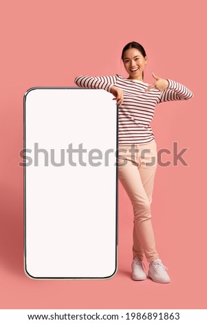 Joyful Asian Lady Leaning And Pointing At Big Smartphone With Blank White Screen, Demonstrating Copy Space For Your App Or Website Design, Standing On Pink Studio Background, Mockup Image Royalty-Free Stock Photo #1986891863