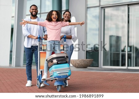 Family Trip Concept. Portrait of cheerful African American girl having fun and spreading hands, ready for vacation, standing on luggage cart. Parents walking with baggage trolley, riding daughter Royalty-Free Stock Photo #1986891899