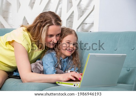 Portrait of mother with her preschool-aged caucasian daughter lying on the couch with a laptop and look at the monitor with a smile. Online learning concept, spending time with children at home.