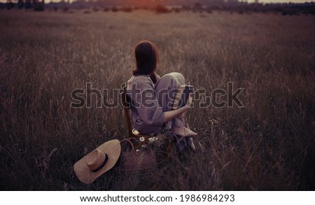 Beautiful woman in linen dress sitting on rustic chair and dreaming in summer meadow. Young female relaxing with book and basket of flowers in countryside. Atmospheric stylish dreamy image Royalty-Free Stock Photo #1986984293