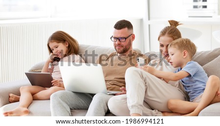 Young happy family, parents with two kids using modern technologies while spending time together at home. Father, mother and children with laptop, digital tablet and smartphone relaxing on sofa