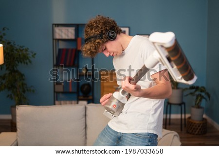 Portrait of a men having fun in the evening while cleaning. A teenager holds a vacuum cleaner like a guitar and dances with it, pretending to play while listening to music on headphones.