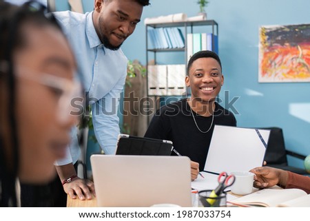 A dark-skinned young boy looks into the camera, smiles as he fills out company reports, signs off on documents, projects; next to him, a senior employee supervises co-workers in the office
