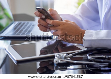 Doctor hand holding and using mobile smart phone and work on laptop computer with digital tablet and medical stethoscope on the table at office in hospital. Medical technology, telehealth concept.